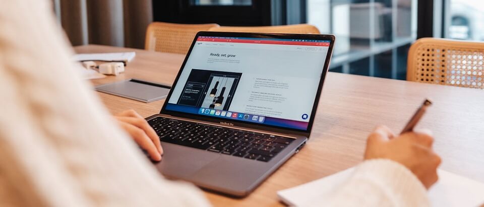 Ein Laptop steht aufgeklappt auf einen Tisch und eine Studierende der University of Applied Sciences Europe macht sich Notizen zu der Webseite die sie im Laptop sieht